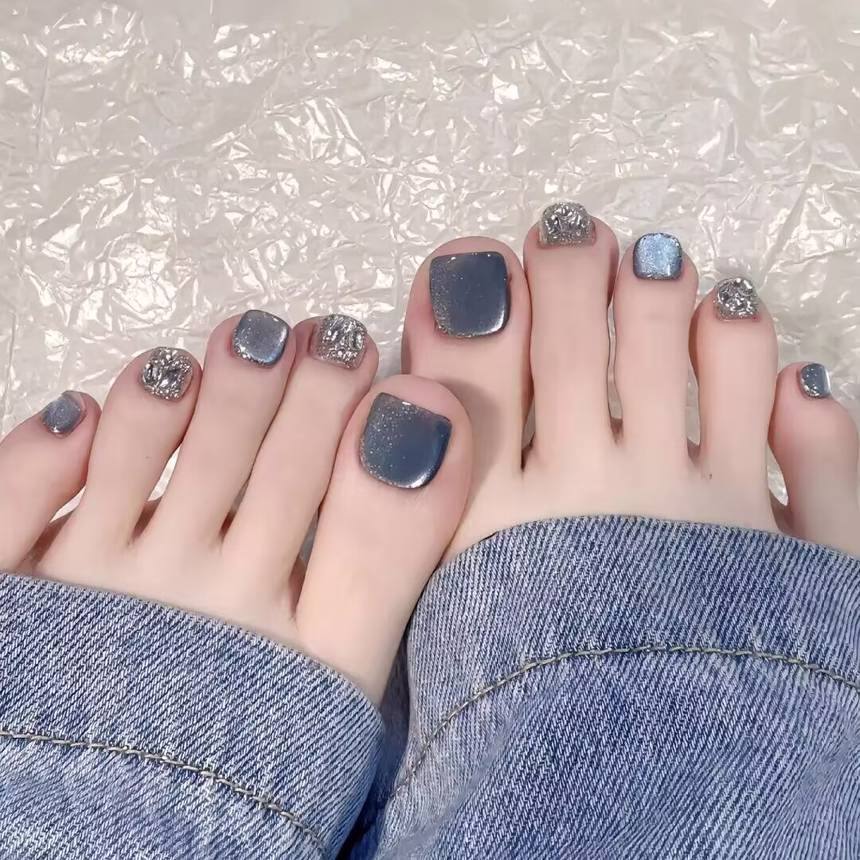 Vibrant summer pedicure trends featuring light blue nail polish with white polka dots on barefoot toes, adorned with a delicate shell anklet, against a sandy shore background.