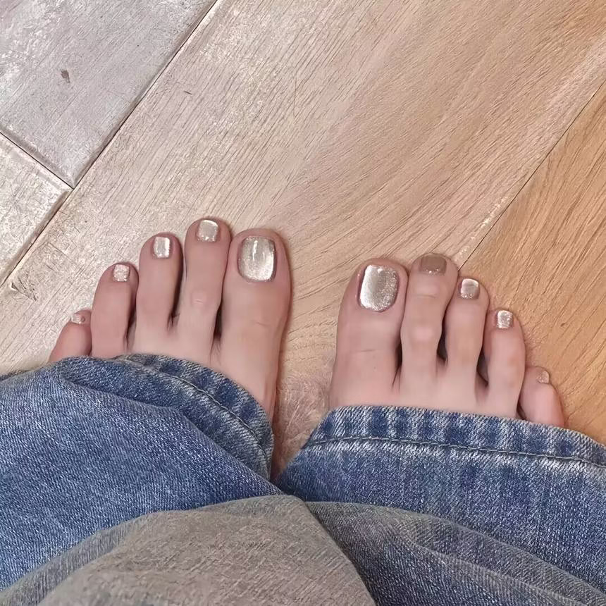 Vibrant summer pedicure trends featuring light blue nail polish with white polka dots on barefoot toes, adorned with a delicate shell anklet, against a sandy shore background.