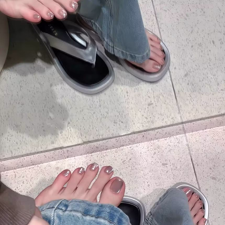 Vibrant summer pedicure trends featuring light blue nail polish with white polka dots on barefoot toes, adorned with a delicate shell anklet, against a sandy shore background.