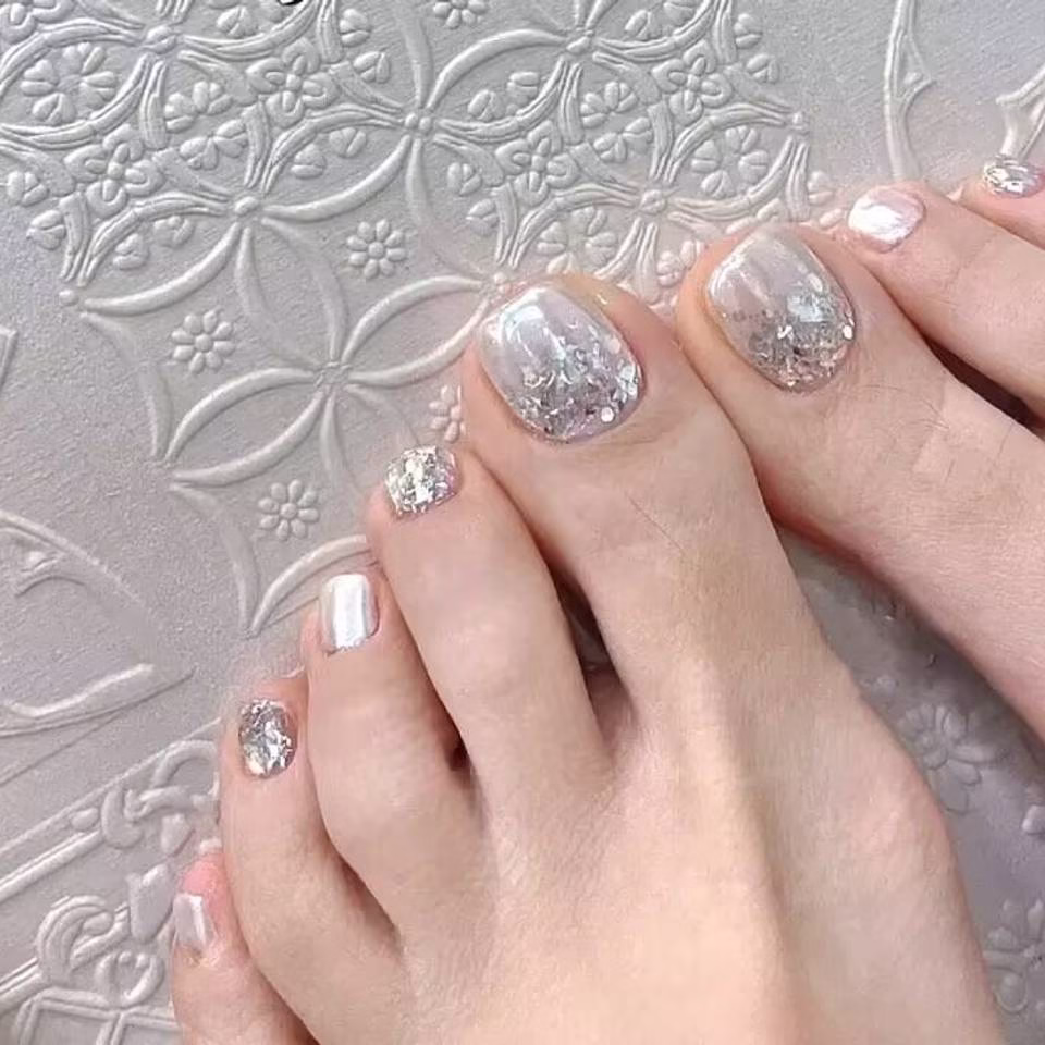 Close-up view of whimsical fairy pedicure designs featuring delicate glitter gradients, tiny floral accents, and ethereal wing motifs on toenails, in soft pastel and iridescent colors.